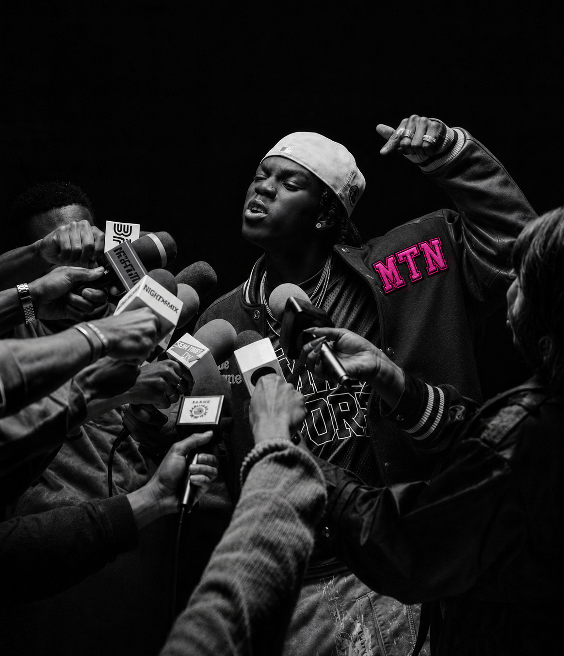 A man wearing a white bandana and a black jacket featuring a pink MTN logo speaking into a cluster of press microphones at a campaign event.