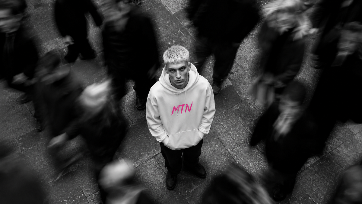 A blurred crowd of people with a single man standing out in the centre, wearing a white hoodie with a pink MTN logo and looking up directly at the camera.