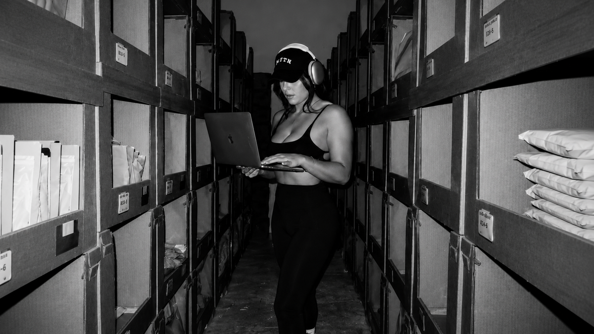 A woman wearing a tank top, hat, and headphones holding a laptop and typing.